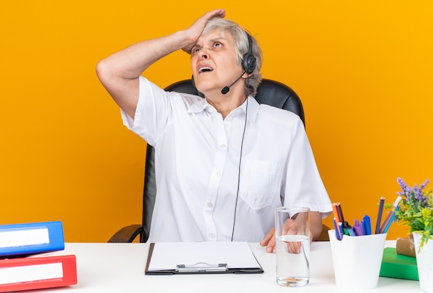 Foto gratuita operatore di call center femminile caucasico scontento sulle cuffie seduto alla scrivania con strumenti da ufficio che mette la mano sulla fronte guardando in alto