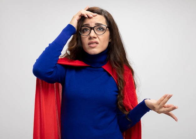 Unpleased caucasian superhero girl with red cape in optical glasses puts hand on forehead on white