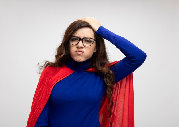 Unpleased caucasian superhero girl with red cape in optical glasses puts hand on head on white