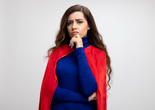 Unpleased caucasian superhero girl with red cape puts hand on chin isolated on white wall with copy space