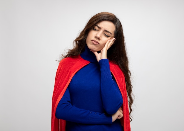 Unpleased caucasian superhero girl with red cape puts hand on face and stands with closed eyes