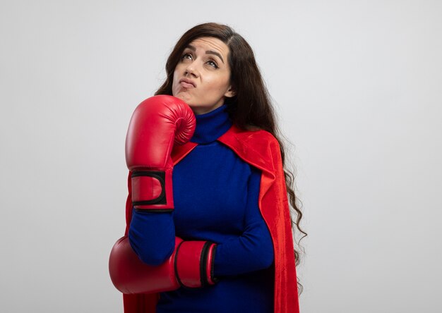 Unpleased caucasian superhero girl with red cape wearing wearing boxing gloves puts fist on chin and looks up isolated on white wall with copy space