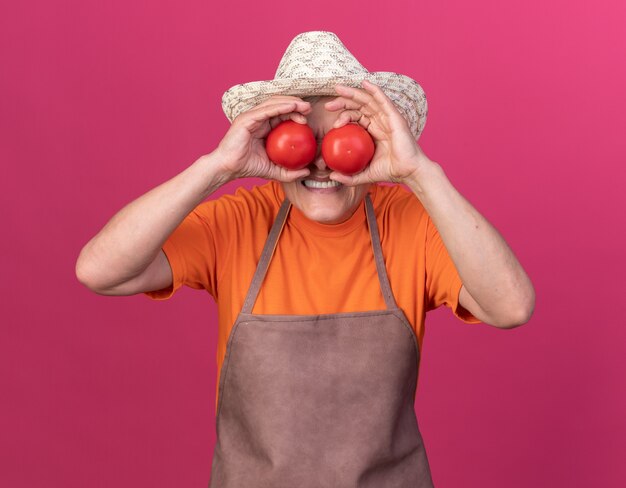 Unpleased elderly female gardener wearing gardening hat covering eyes with tomatoes