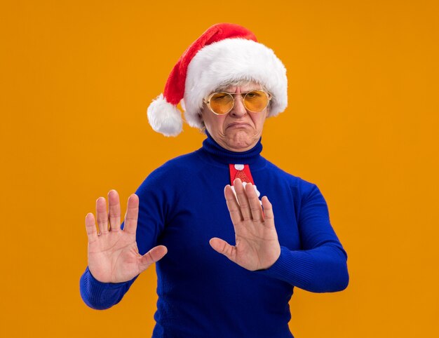 Unpleased elderly woman in sun glasses with santa hat and santa tie holds hands open gesturing no sign isolated on orange wall with copy space