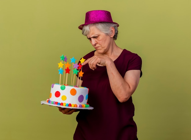 Unpleased elderly woman wearing party hat holds and points at birthday cake isolated on olive green wall with copy space