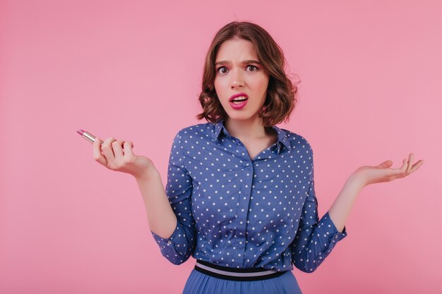 Unpleased female model in trendy blouse posing with lipstick. Charming curly lady expressing negative emotions while doing her makeup.