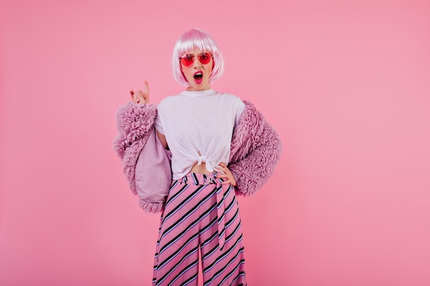 Free photo unpleased female model in trendy fluffy jacket posing . attractive disappointed girl in pink periwig isolated on colorful wall