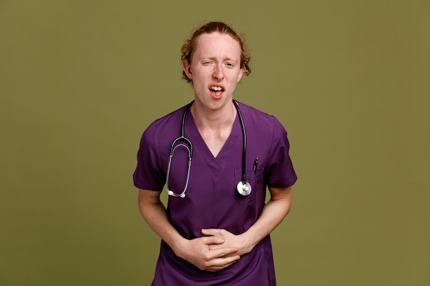 Foto gratuita giovane medico maschio scontento dello stomaco che indossa l'uniforme con lo stetoscopio isolato su sfondo verde