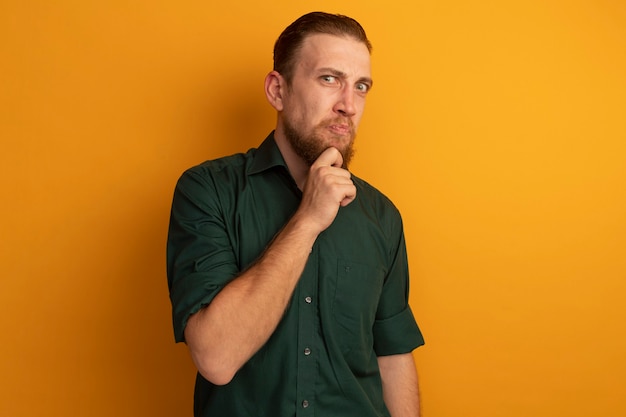Unpleased handsome blonde man holds chin isolated on orange wall