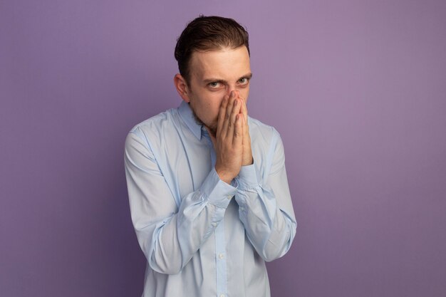 Unpleased handsome blonde man puts hands on nose isolated on purple wall