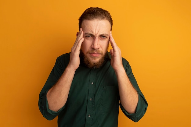 Unpleased handsome blonde man puts hands on temples isolated on orange wall