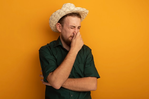 Unpleased handsome blonde man with beach hat closes nose isolated on orange wall