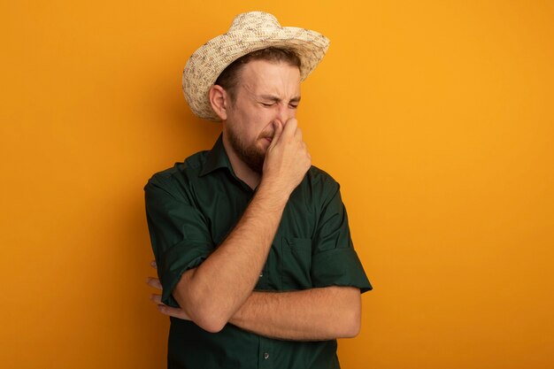 비치 모자와 불쾌한 잘 생긴 금발의 남자가 오렌지 벽에 고립 된 코를 닫습니다.