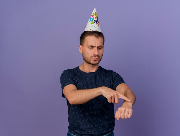 Unpleased handsome caucasian man wearing birthday cap looks and points at hand isolated on purple background with copy space