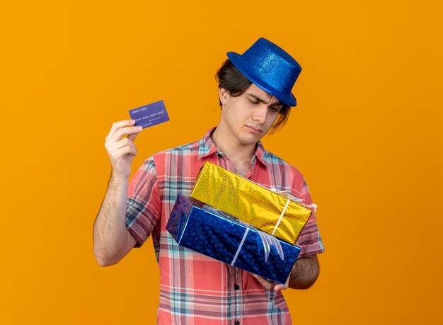 Unpleased handsome caucasian man wearing blue party hat holds gift boxes and credit card 