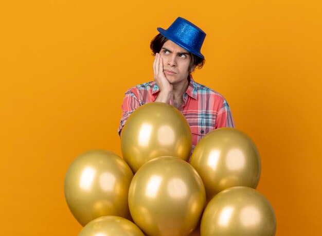 Unpleased handsome caucasian man wearing blue party hat puts hand on face and stands