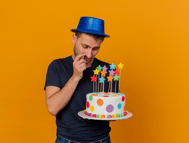 Unpleased handsome man wearing blue party hat holds and looks at birthday cake isolated on orange wall with copy space