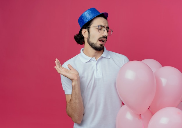 Недовольный красавец в очках и синей шляпе держит и смотрит на воздушные шары и протягивает руку, изолированную от розового