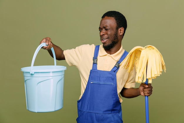 Unpleased holding bucket with mop young africanamerican cleaner male in uniform with gloves isolated on green background