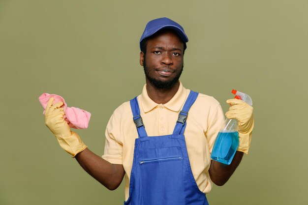 Unpleased holding cleaning agent with rag young africanamerican cleaner male in uniform with gloves isolated on green background