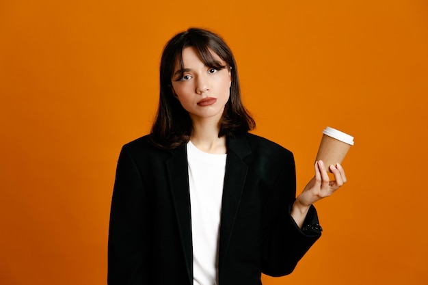 Недовольная держа чашку кофе молодая красивая женщина в черной куртке на оранжевом фоне