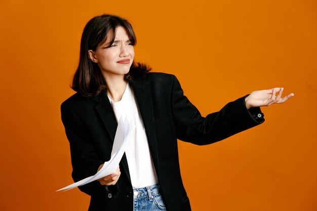 オレンジ色の背景に分離された黒いジャケットを着ている若い美しい女性の不愉快な保持紙