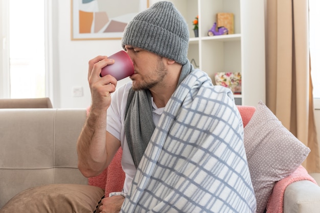 Unpleased ill slavic man with scarf around neck wearing winter hat wrapped in plaid drinking from cup sitting on couch at living room