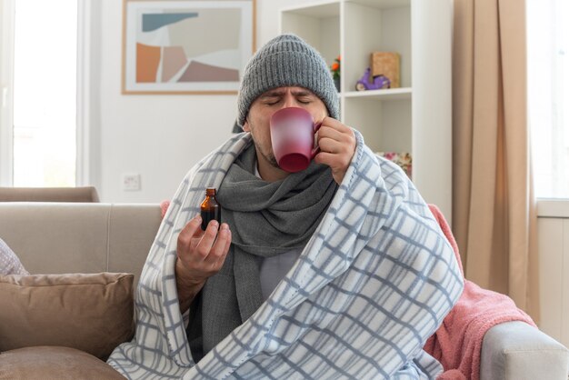 Unpleased ill slavic man with scarf around neck wearing winter hat wrapped in plaid drinking medicine from cup sitting on couch at living room