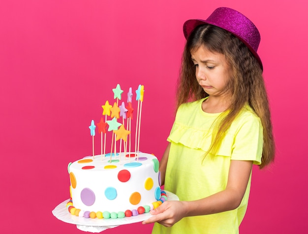 unpleased little caucasian girl with purple party hat holding and looking at birthday cake isolated on pink wall with copy space