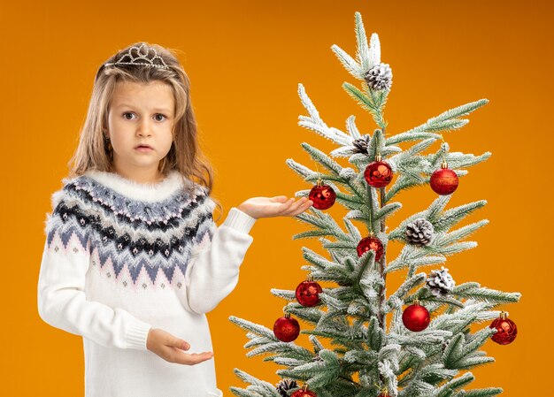 Unpleased little girl standing nearby christmas tree wearing tiara with garland on neck points at side isolated on orange background