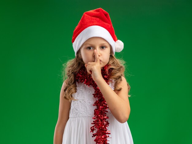 Unpleased little girl wearing christmas hat with garland on neck showing silence gesture isolated on green background