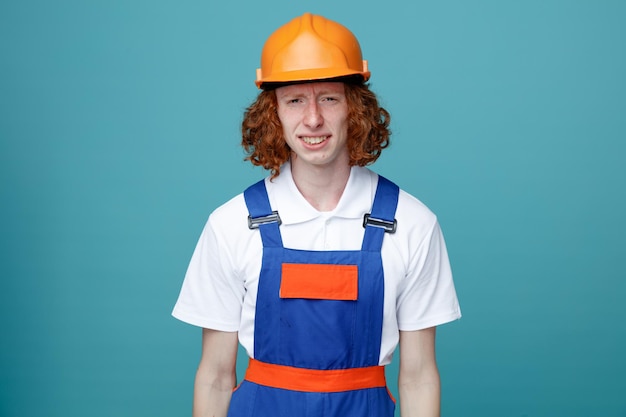 Бесплатное фото Недовольный глядя в камеру молодой строитель в форме на синем фоне