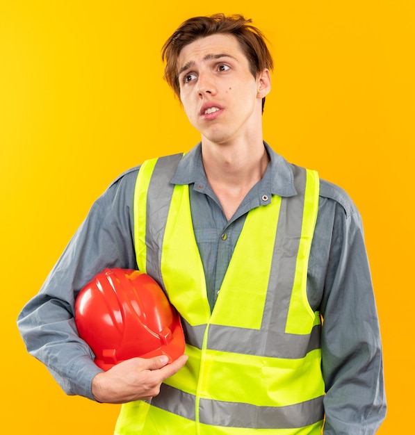 Unpleased looking atside young builder man in uniform holding safety helmet 