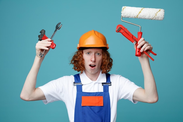Unpleased looking at camera young builder man in uniform holding construction tools isolated on blue background