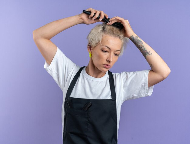 Unpleased looking down young beautiful female barber in uniform combing hair isolated on blue background