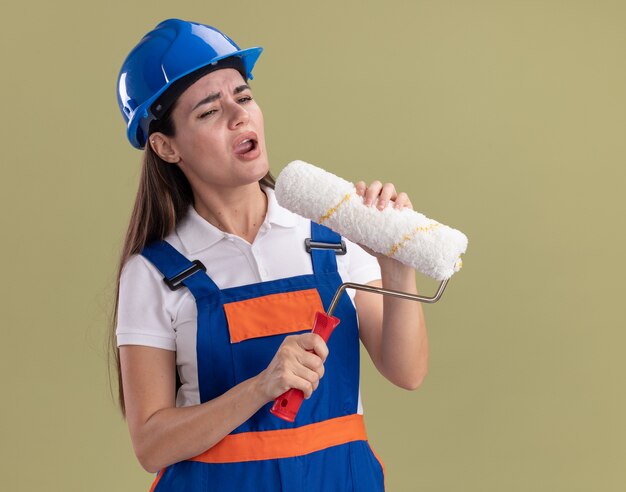 Unpleased looking at side young builder woman in uniform holding roller brush isolated on olive green wall