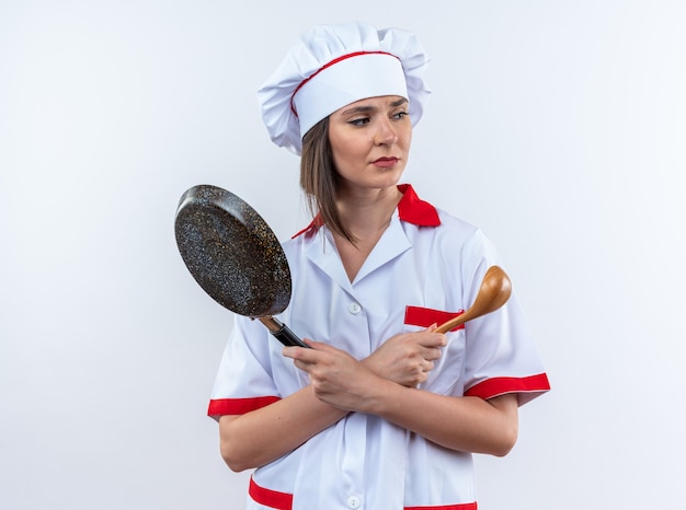 unpleased looking side young female cook wearing chef uniform holding and crossing frying pan with spoon isolated on white wall