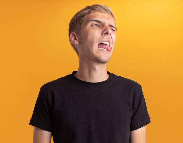 Unpleased looking at side young handsome guy wearing black shirt showing tongue isolated on yellow wall