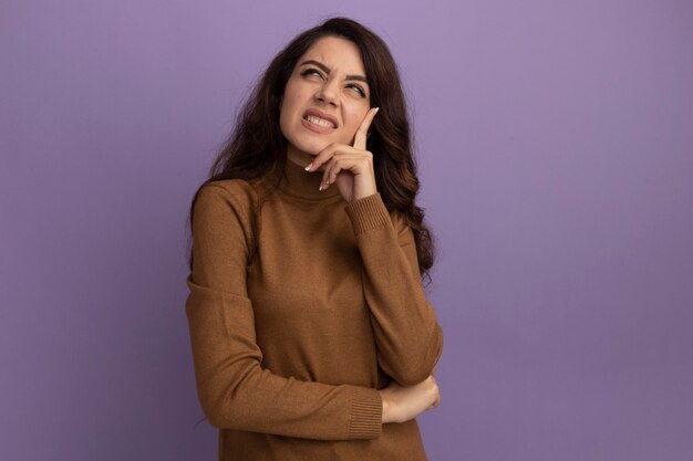 Unpleased looking up young beautiful girl wearing brown turtleneck sweater putting hand on chin isolated on purple wall