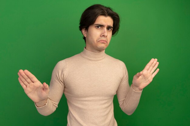 Unpleased looking  young handsome guy showing stop gesture isolated on green wall