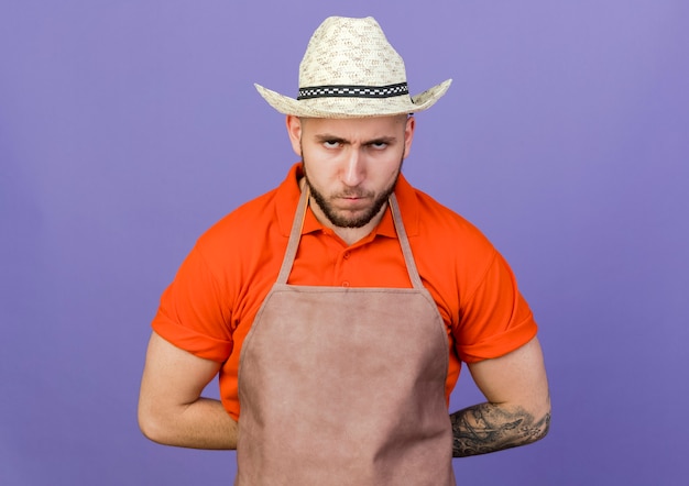 Il giardiniere maschio scontento che porta il cappello di giardinaggio esamina la macchina fotografica