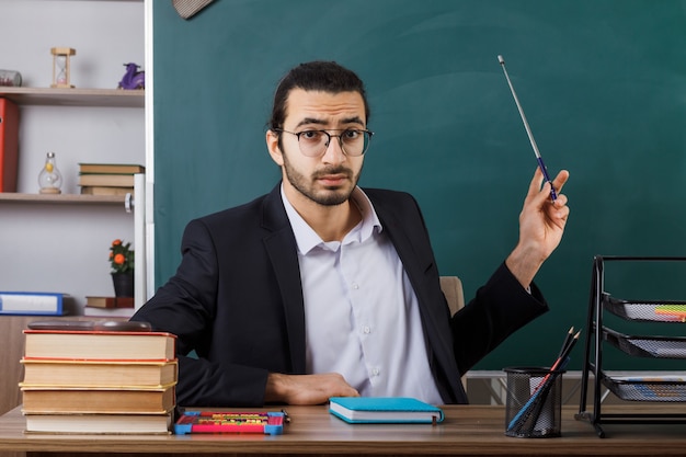 Free photo unpleased male teacher wearing glasses points with pointer stick at blackboard sitting at table with school tools in classroom