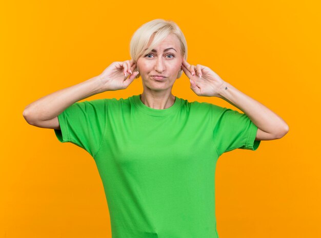 Unpleased middle-aged blonde slavic woman looking at camera keeping fingers on ears isolated on yellow background