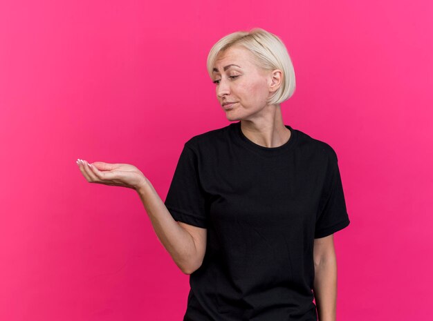 Unpleased middle-aged blonde slavic woman showing empty hand looking at it isolated on pink wall