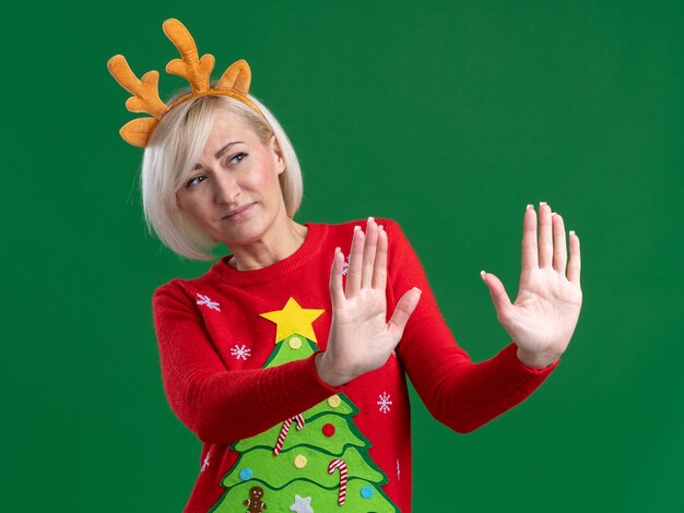 Unpleased middle-aged blonde woman wearing christmas reindeer antlers headband and christmas sweater looking at side doing refusal gesture isolated on green background