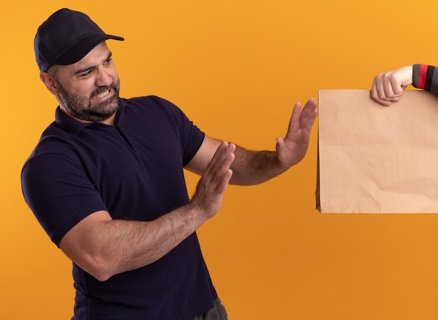 노란색 벽에 고립 된 클라이언트에게 종이 음식 패키지를주는 유니폼과 모자에 불쾌한 중년 배달 남자