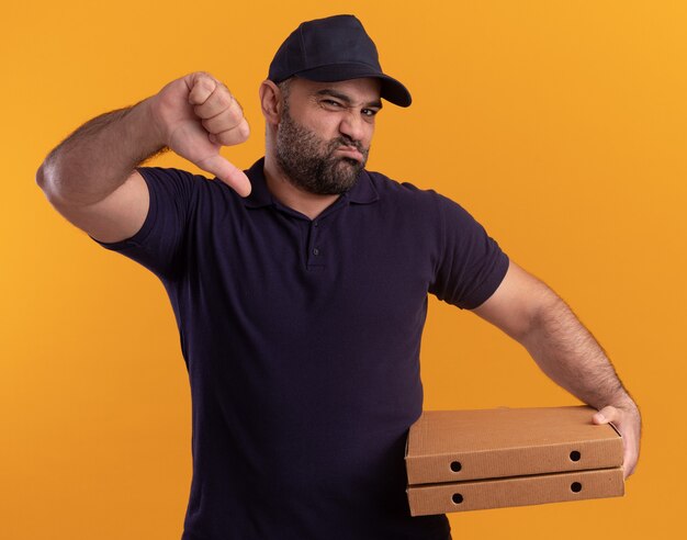 Unpleased middle-aged delivery man in uniform and cap holding pizza boxes showing thumb down isolated on yellow wall