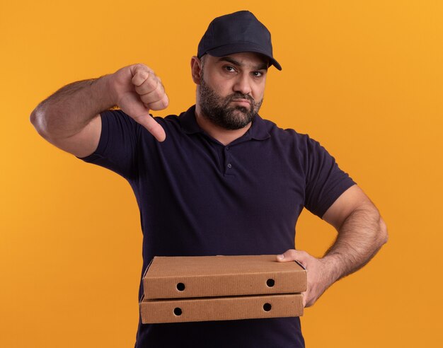 Unpleased middle-aged delivery man in uniform and cap holding pizza boxes showing thumb down isolated on yellow wall