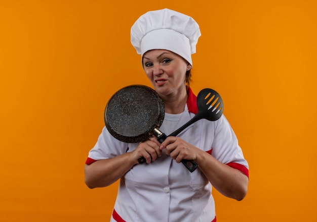 Unpleased middle-aged female cook in chef uniform crossing frying pan and spatula in her hands 
