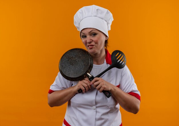 Недовольная женщина-повар средних лет в униформе шеф-повара скрещивает сковороду и лопатку в руках